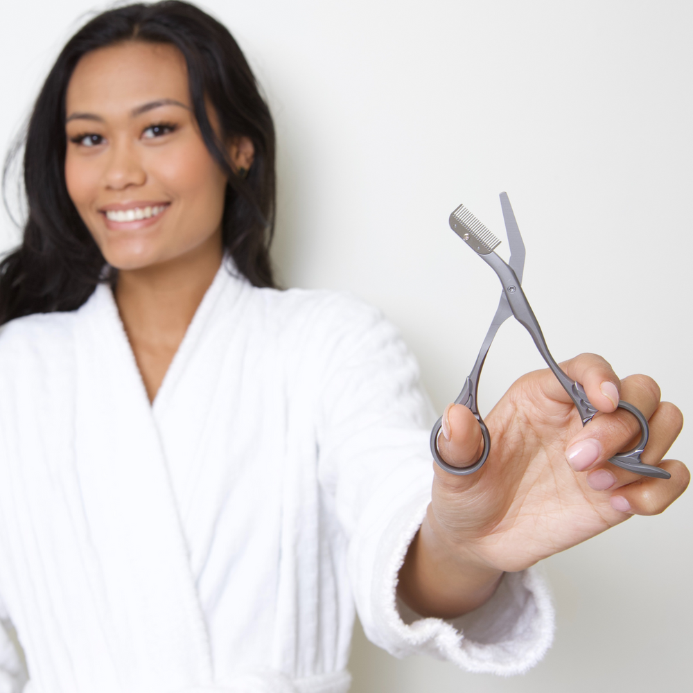 Woman in a white robe holding a pair of scissors and tweezers against a plain background
