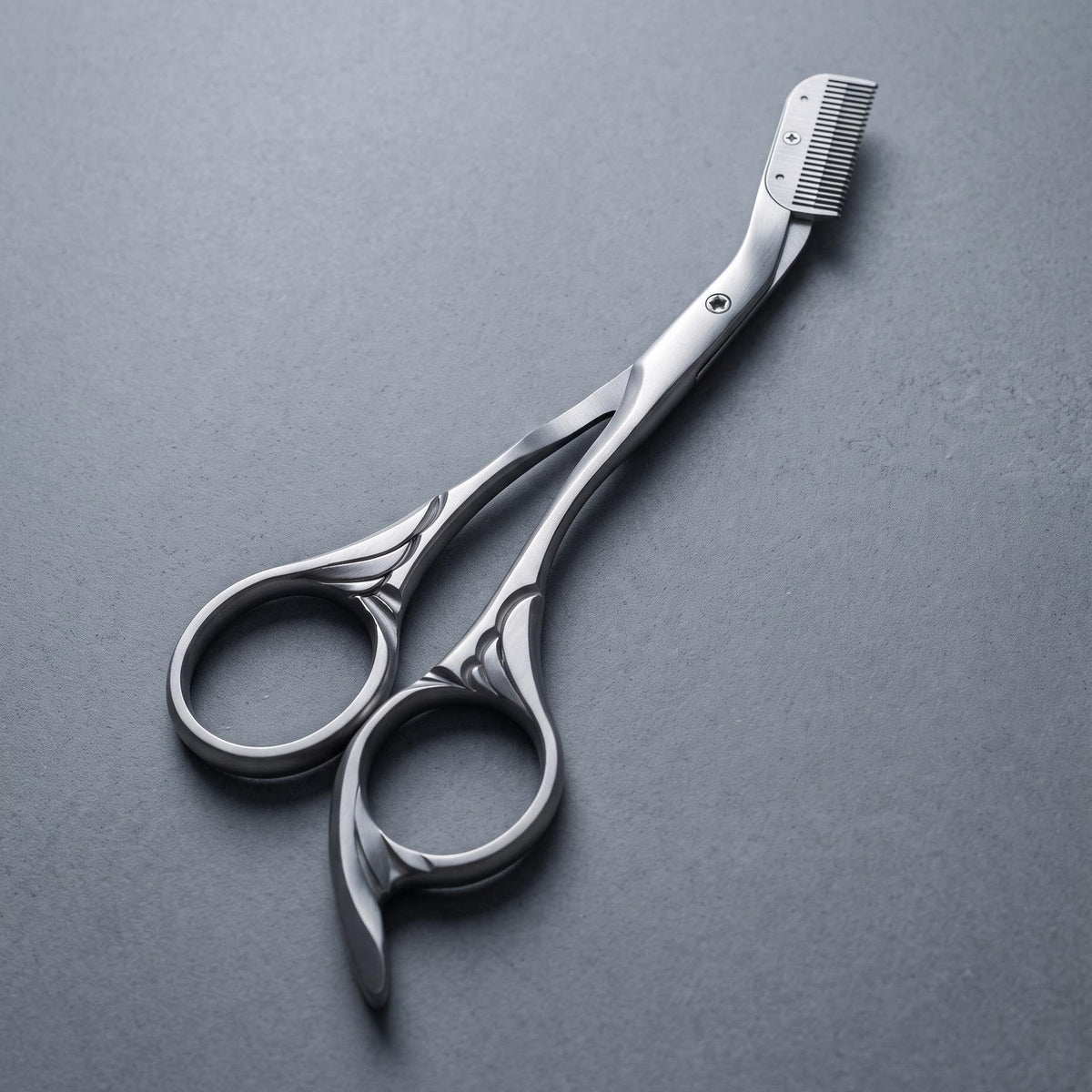 A pair of silver professional eyebrow styling scissors with a built-in eyebrow comb attachment, displayed diagonally on a textured dark gray background.